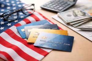 The image presents a clean and realistic composition featuring several credit cards placed over the United States flag, alongside dollar bills and office items. The balanced lighting and minimal visual elements create a professional atmosphere that evokes financial decision-making and economic structure.